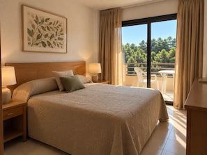 A hotel room with a large bed, two bedside lamps, a framed artwork on the wall, and a balcony with a view of trees. - Costa Brava Beach Apartment for 4 (Pals)