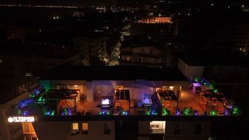 En bygning med flere etager og en tagterrasse med udendørs siddepladser og stemningsbelysning.