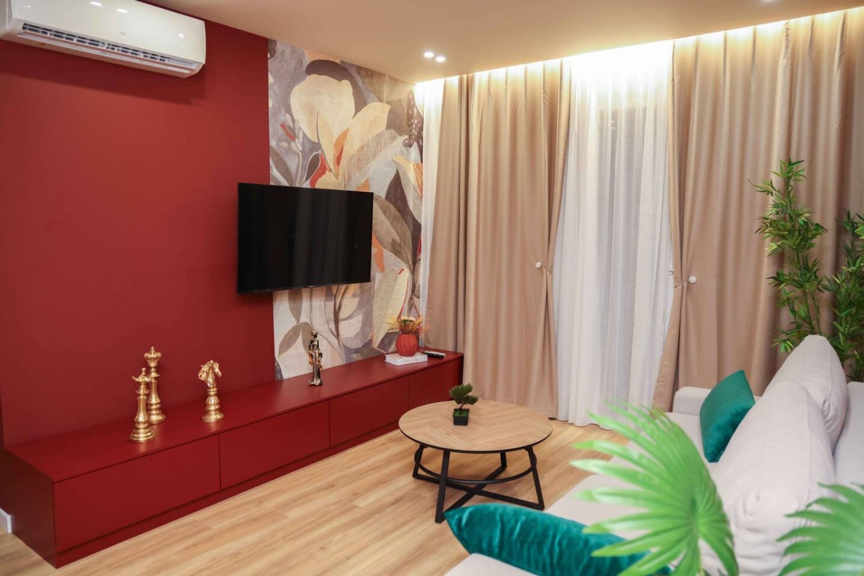 A modern living room with a red accent wall, a mounted TV, a small round table, and a sofa with teal cushions.