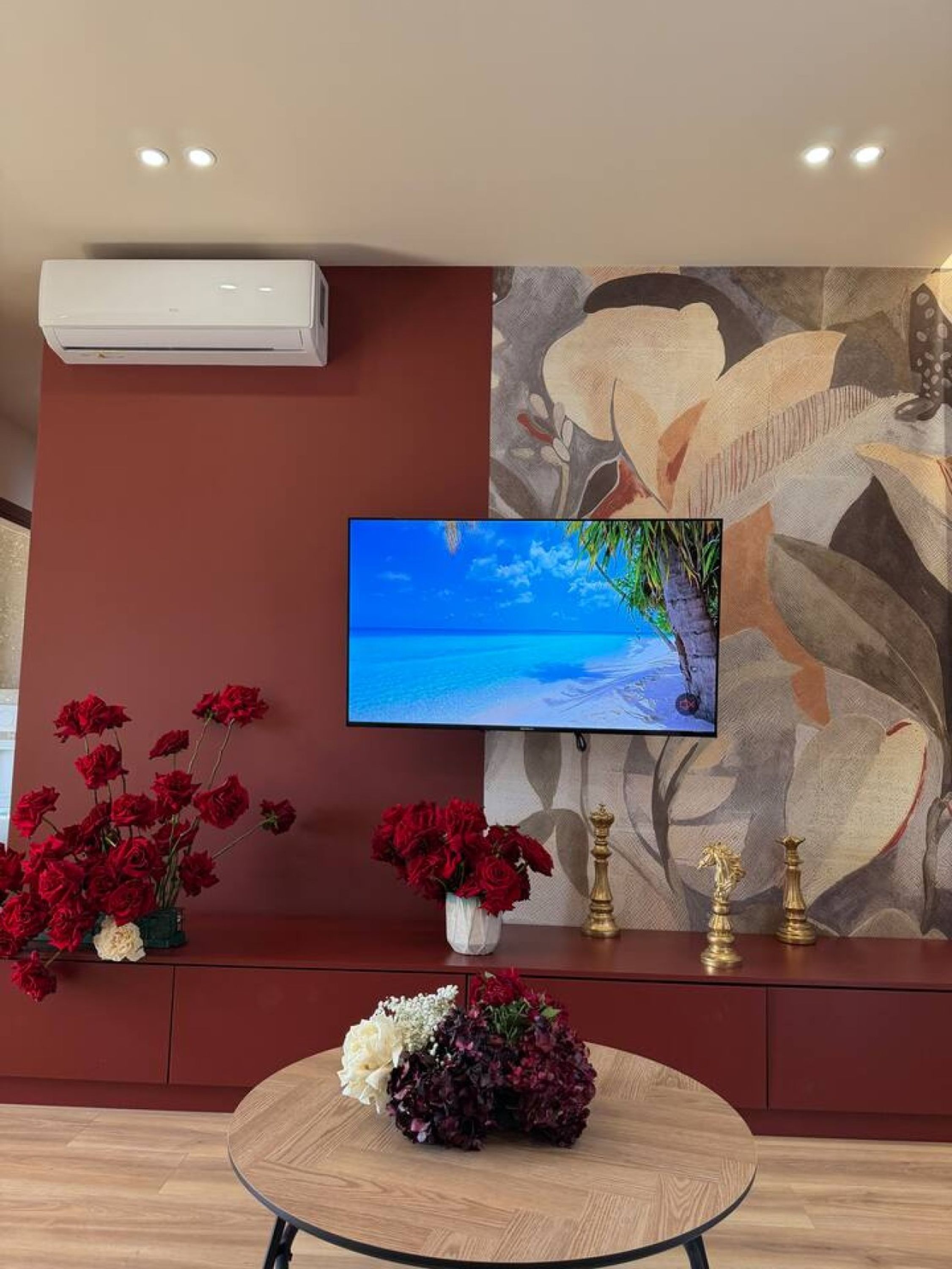 A room with a flat-screen TV mounted on the wall, a red sofa, and a round wooden table with floral arrangements.