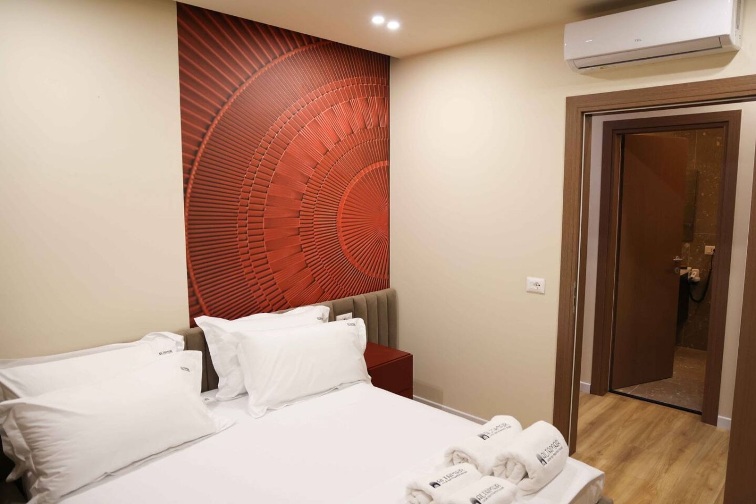 A hotel room with a bed, a red textured wall, an air conditioning unit, and a door leading to a bathroom.
