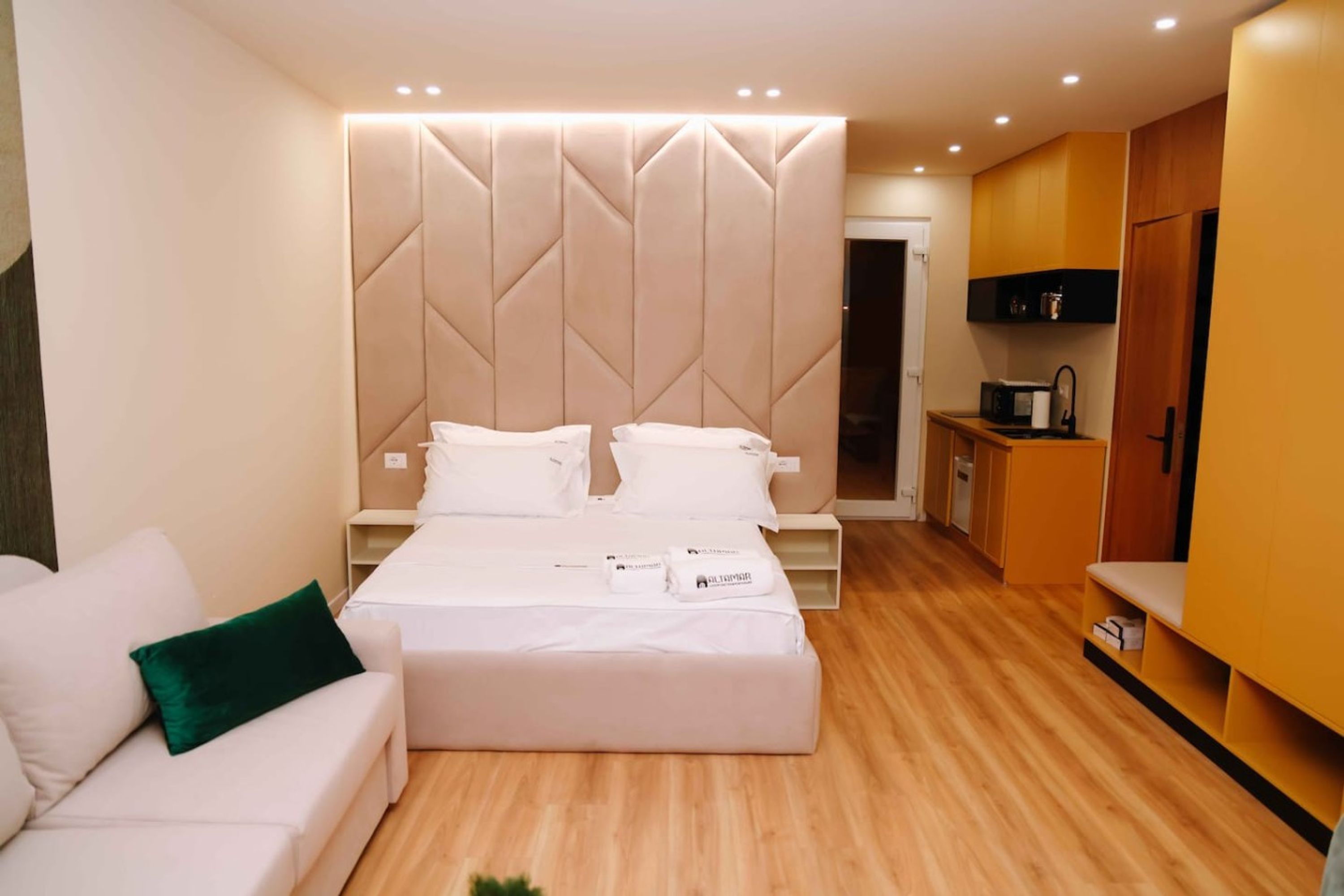 A modern hotel room with a bed, a sofa, a kitchenette, and a bathroom.