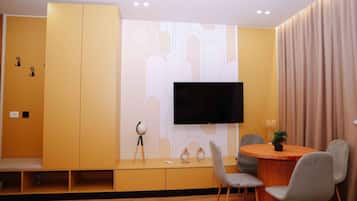 A modern living room with a flat-screen TV mounted on the wall, a wooden bench, a dining table with chairs, and a small potted plant.