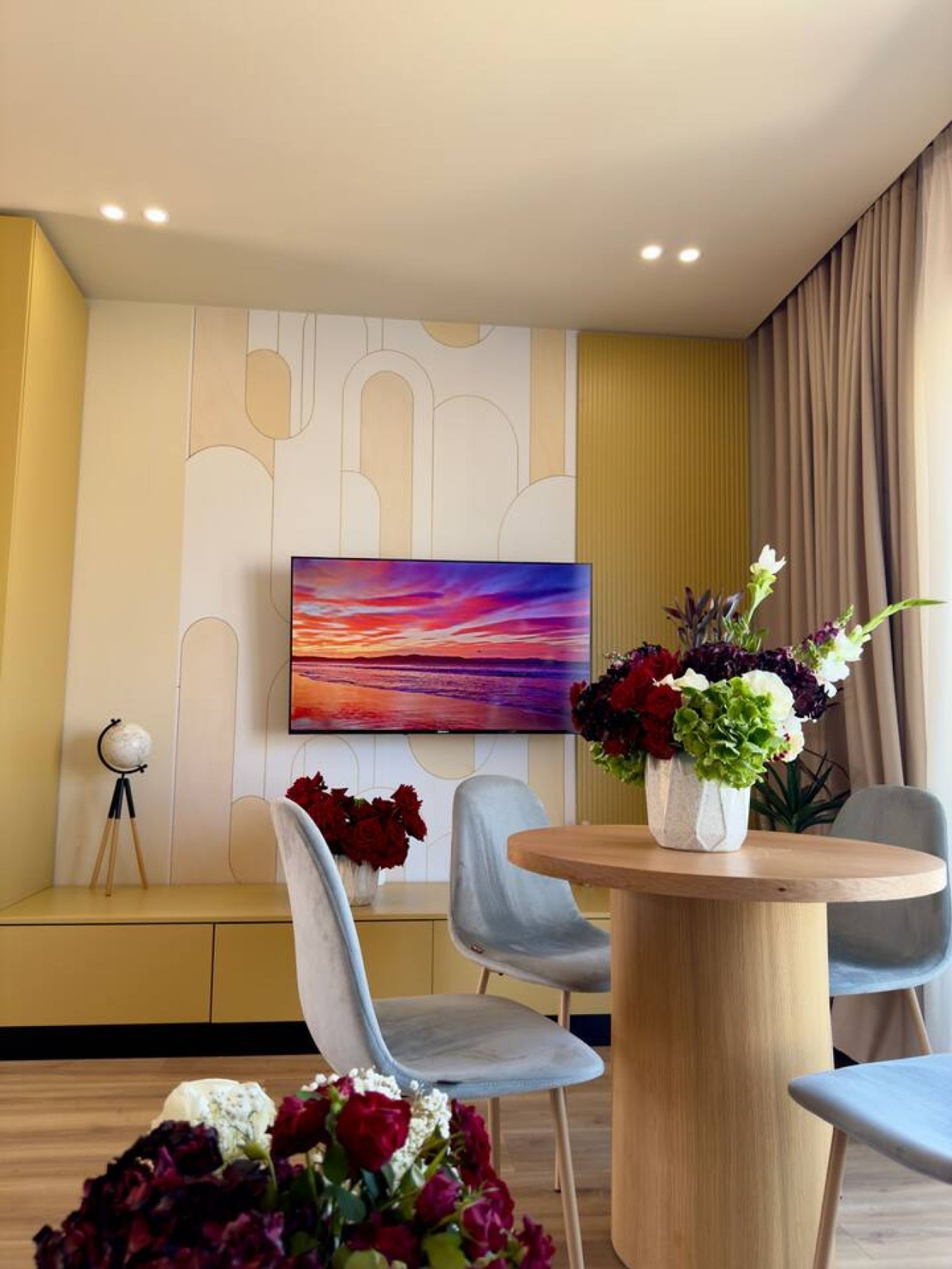 Una habitación moderna con una mesa redonda de madera, dos sillas grises, un televisor de pantalla plana montado en la pared y un muro decorativo con patrones geométricos.