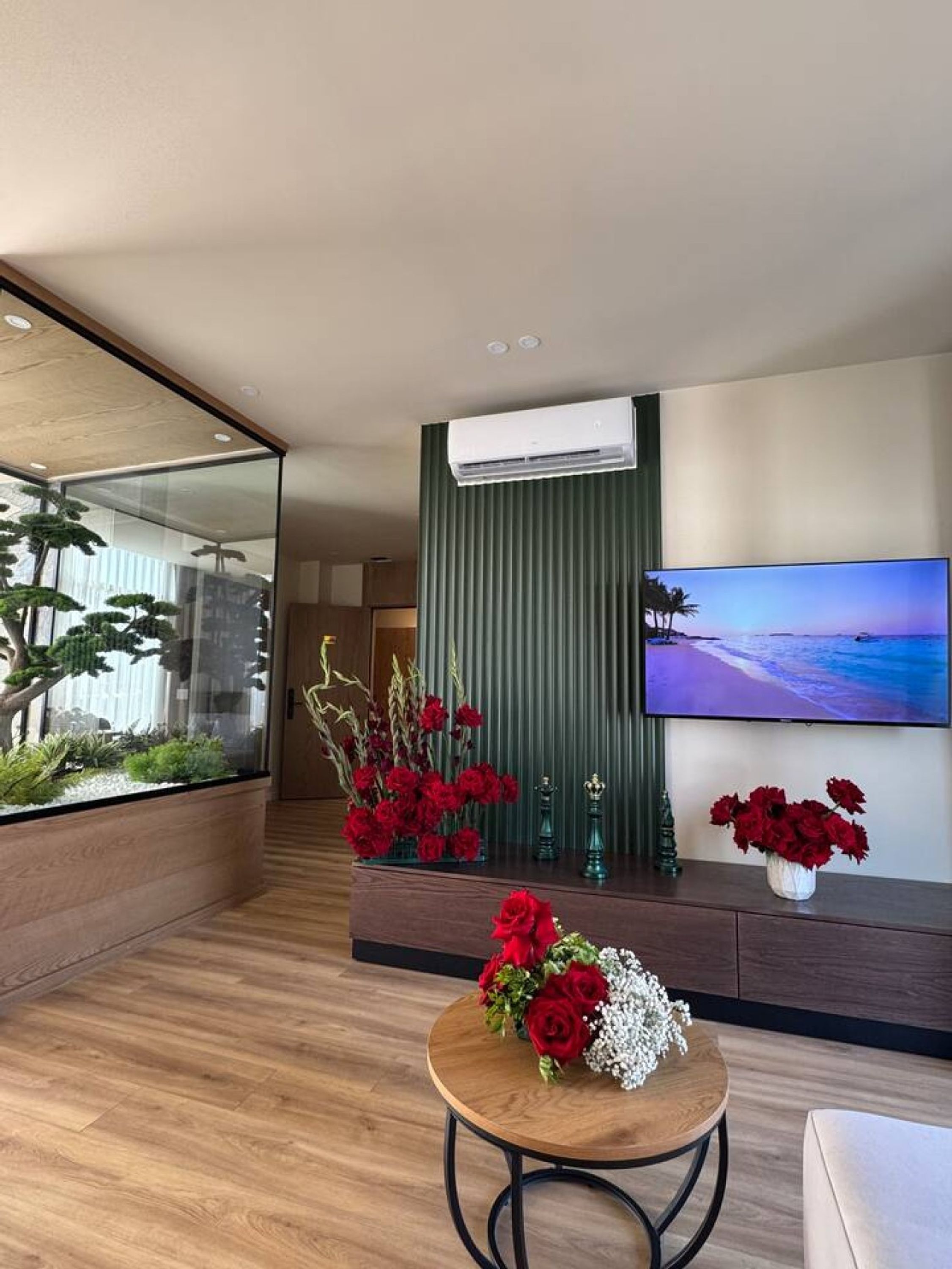 A modern living room with a glass partition, a mounted air conditioner, a flat-screen TV, and a wooden coffee table with a floral arrangement.