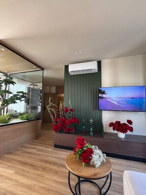 Ein modernes Wohnzimmer mit Glastrennwand, einer Wandklimaanlage, einem Flachbildfernseher und einem Couchtisch mit floralem Arrangement.
