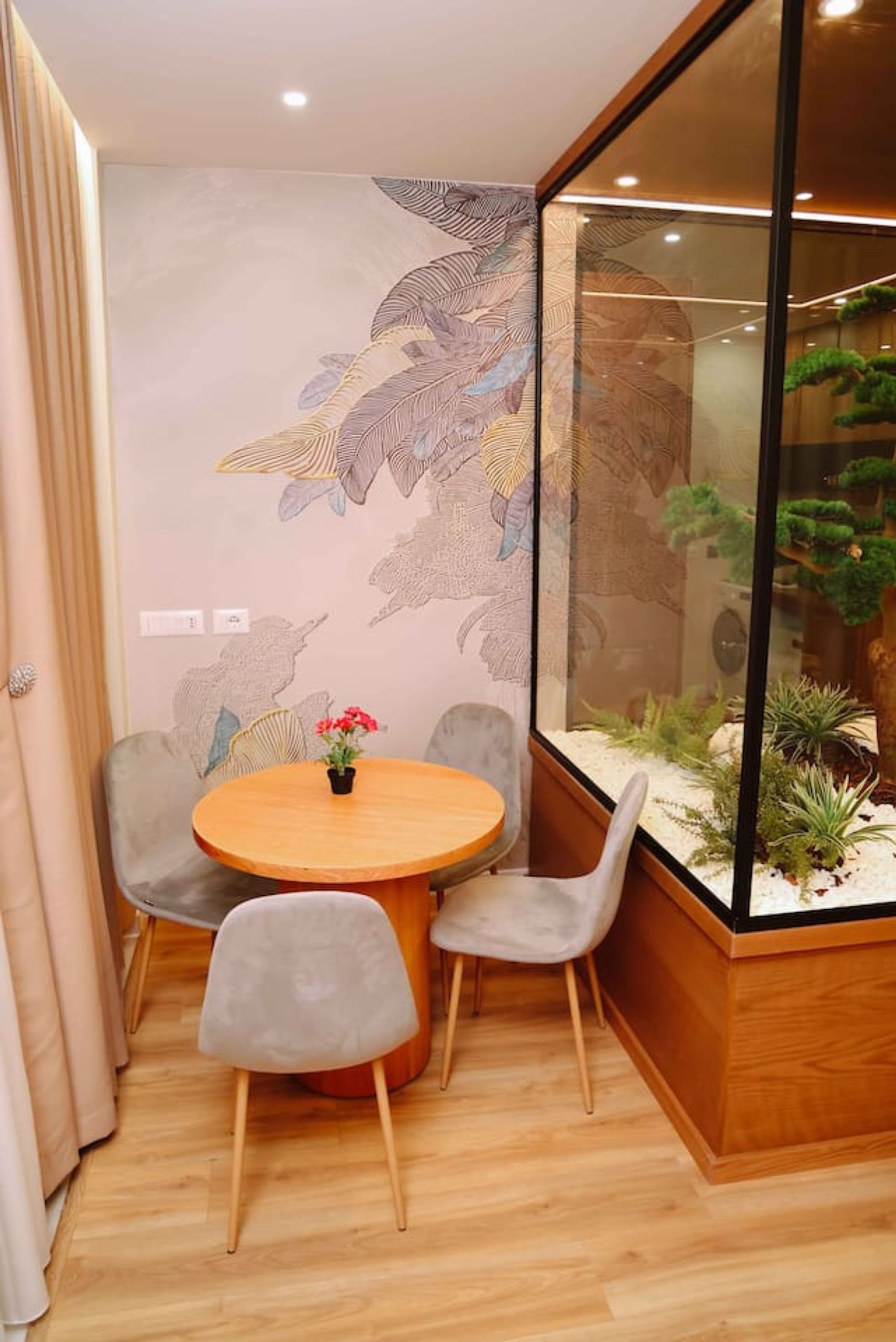 A room with a round wooden table, two grey chairs, a wall with a leaf pattern, and a glass enclosure with plants.