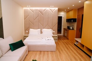 Una habitación de hotel moderna con una cama grande, una mesita de noche, un escritorio pequeño, una kitchenette y un armario.