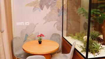 A room with a round wooden table, two grey chairs, a decorative wall with a leaf pattern, and a glass enclosure with a plant.
