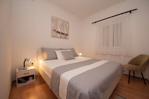 A bedroom with a bed, nightstand, chair, and artwork on the wall. - Spacious apartment with balcony, big garden & grill, near beach (Podglavica)