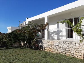 A modern white building with a stone wall foundation, a covered porch, and a tree with fruit. - Spacious apartment with balcony, big garden & grill, near beach (Podglavica)