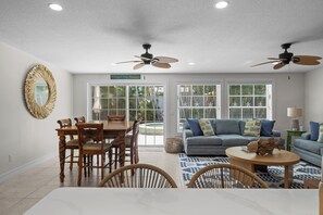 A spacious living area with a dining table, chairs, a sofa, and a coffee table. There are ceiling fans, a large mirror, and a view of a pool through the glass doors. - Lucky 307- 3 bedroom with pool and spa- Managed by Beach House Real Estate (Holmes Beach)