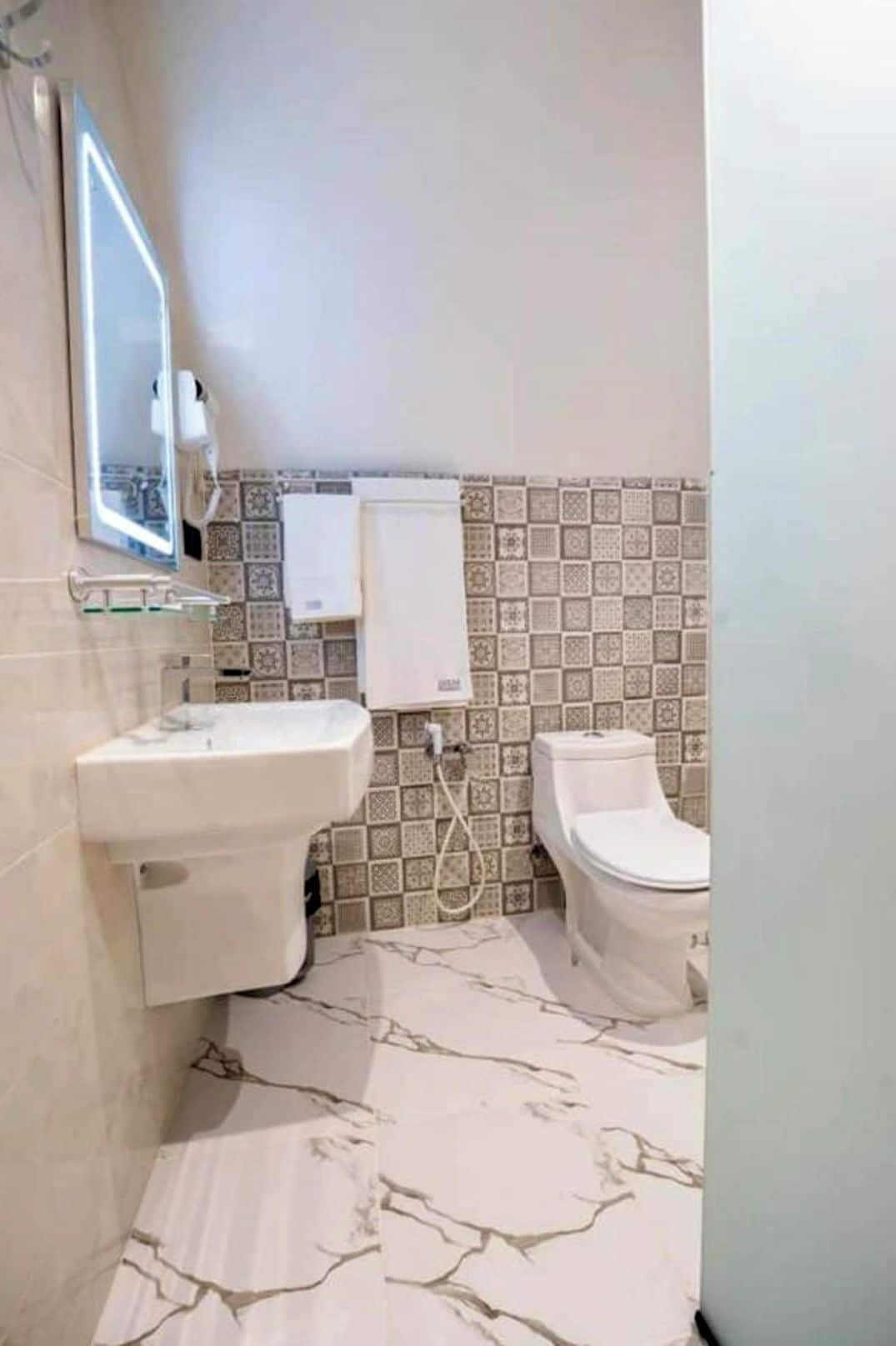 Un baño con un lavamanos blanco, un inodoro y un espejo. El suelo presenta un diseño de mármol.
