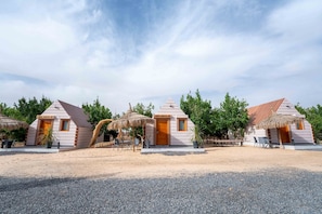 Three thatched-roof cabins with wooden doors, arranged in a row on a gravel surface, surrounded by greenery.