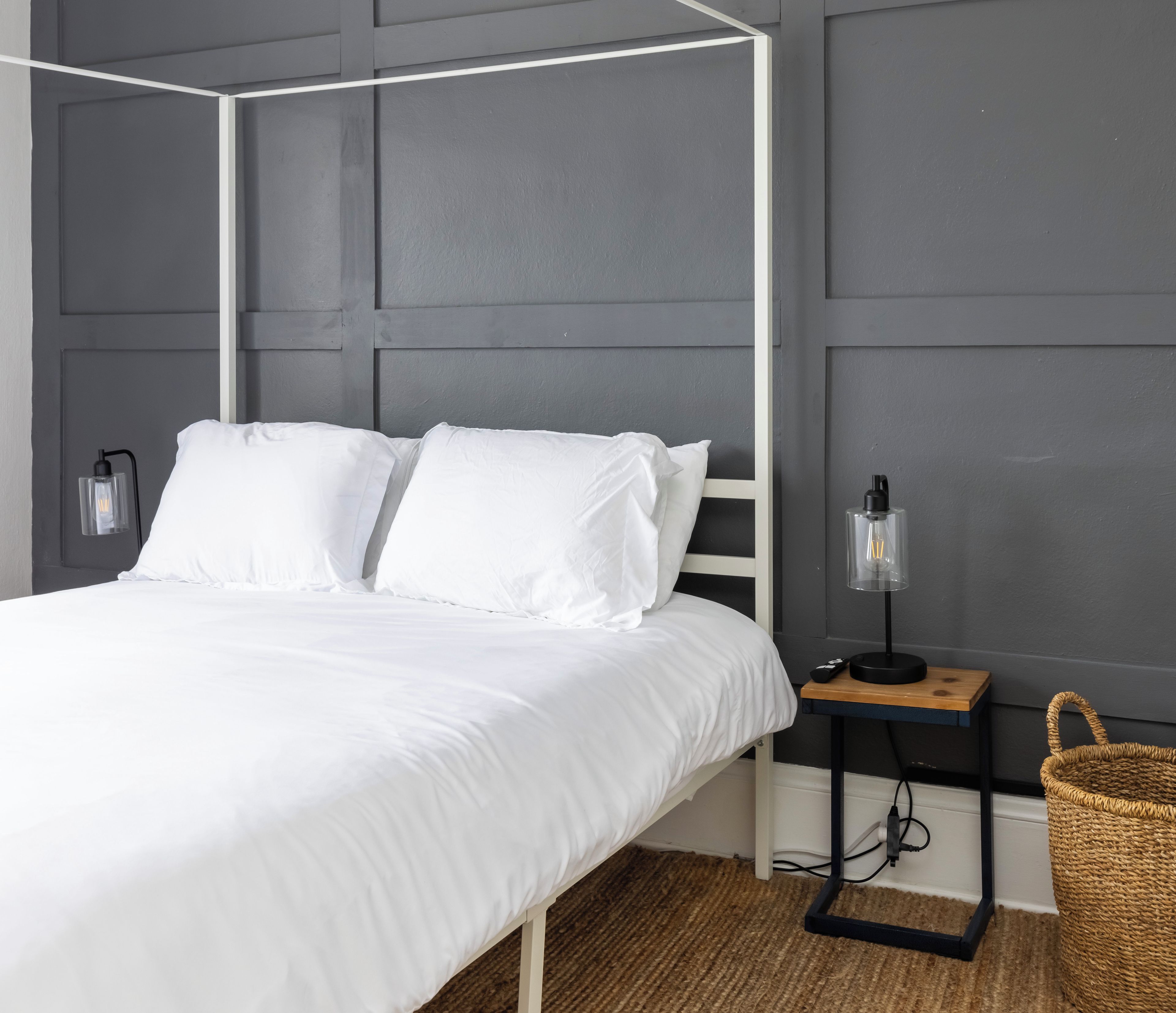 A bedroom with a four-poster bed, two bedside lamps, a bedside table with a lamp, and a woven basket.