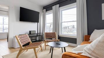 A modern living room with a flat-screen TV mounted on the wall, two chairs with woven backs, a round coffee table, and a sofa with a white throw.