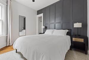 Un dormitorio moderno con una cama grande, una mesita de noche, un tocador, un espejo y una ventana con cortinas.
