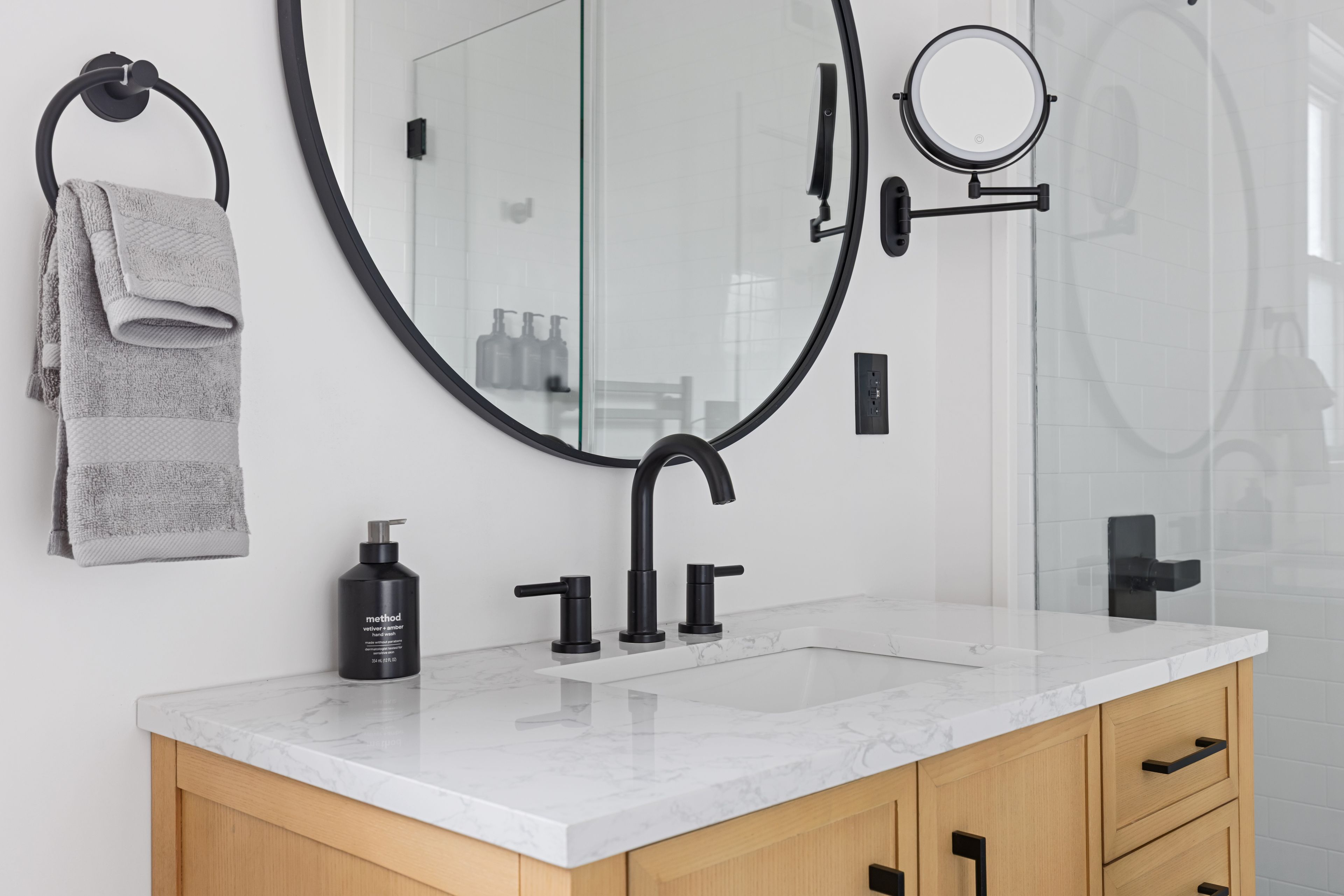 A bathroom with a large mirror, a round mirror, a towel rack, a sink, and a bottle of hand wash.