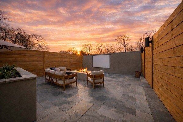 Un patio al aire libre con una chimenea, dos sillas de madera y una pantalla grande.