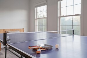 A ping pong table with a paddle, books, and a ball in a room with large windows. - Lake Views, Soaking Tub, Ping Pong Table, Firepit (Sharps Chapel)