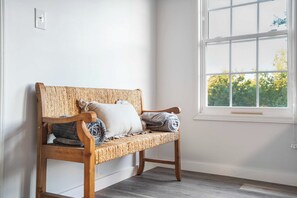 A wooden bench with woven backrest and cushions against a white wall, next to a window with a view of trees. - Lake Views, Soaking Tub, Ping Pong Table, Firepit (Sharps Chapel)