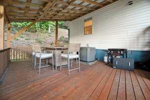 A covered outdoor area with a wooden deck, two metal chairs with cushions, a table, and a grill unit. - Lake Views, Soaking Tub, Ping Pong Table, Firepit (Sharps Chapel)