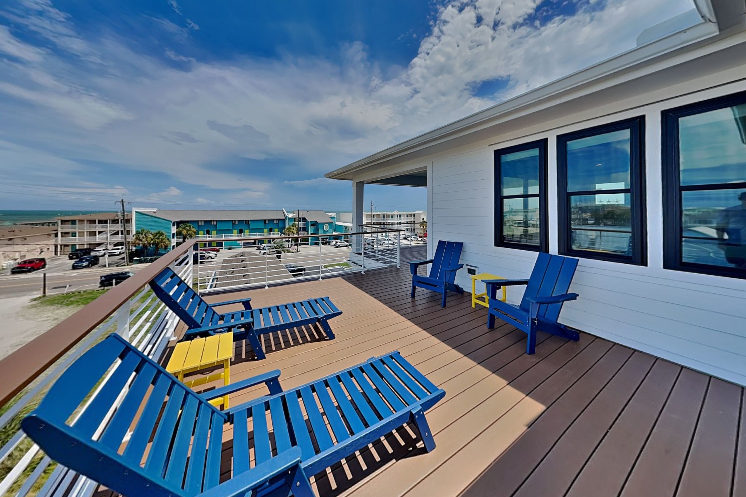 A deck with blue and yellow lounge chairs, a small table, and a view of the beach and buildings in the background.