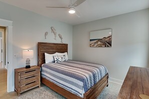 A bedroom with a wooden bed, a nightstand, a ceiling fan, a wall-mounted painting, and a herringbone-patterned rug. - Klose Enough (Kure Beach)
