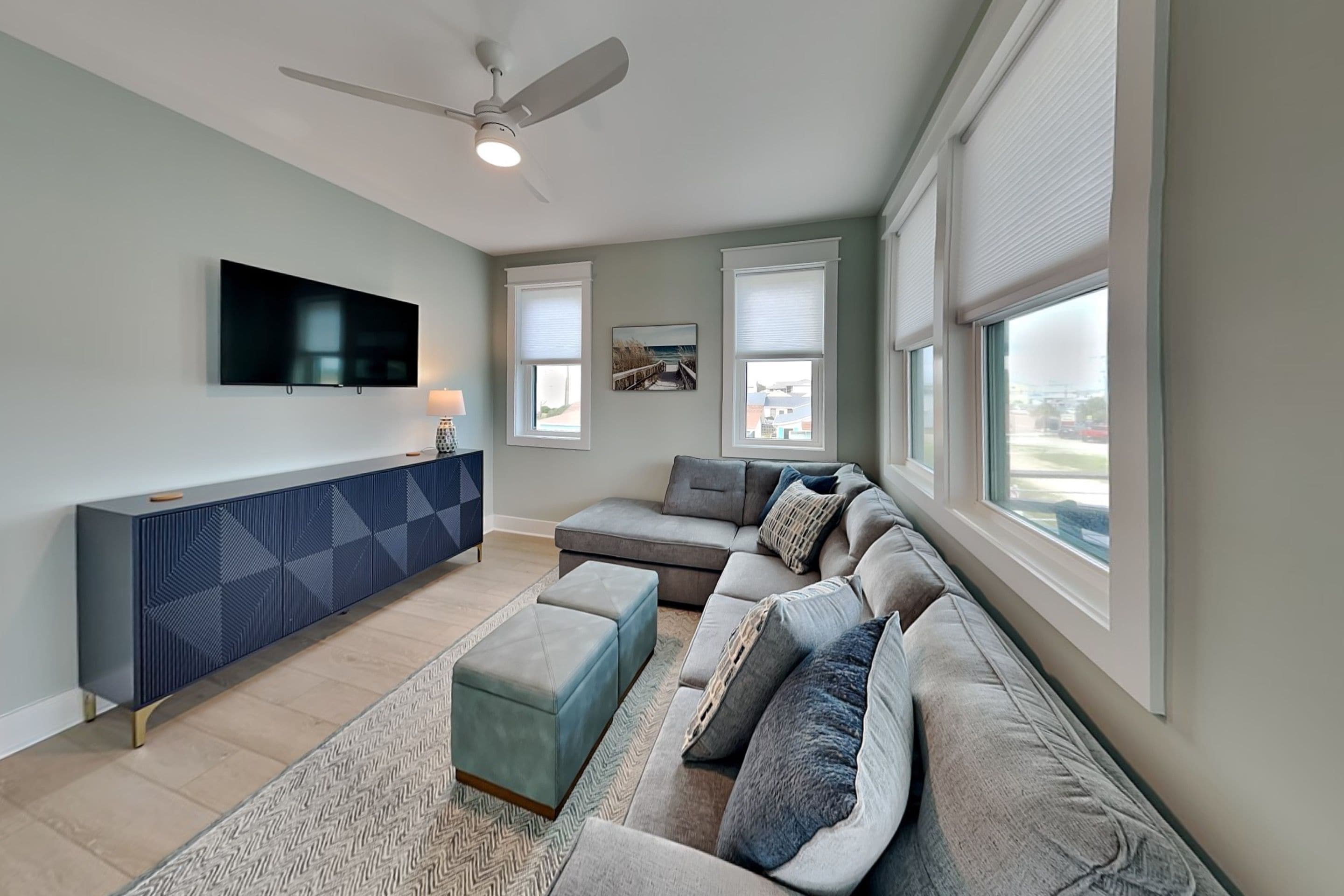 A living room with a sectional sofa, a flat-screen TV, a ceiling fan, and large windows.