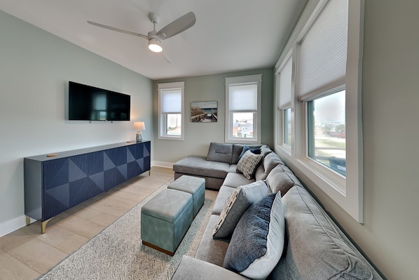 A living room with a sectional sofa, a flat-screen TV, a ceiling fan, and large windows. - Klose Enough (Kure Beach)