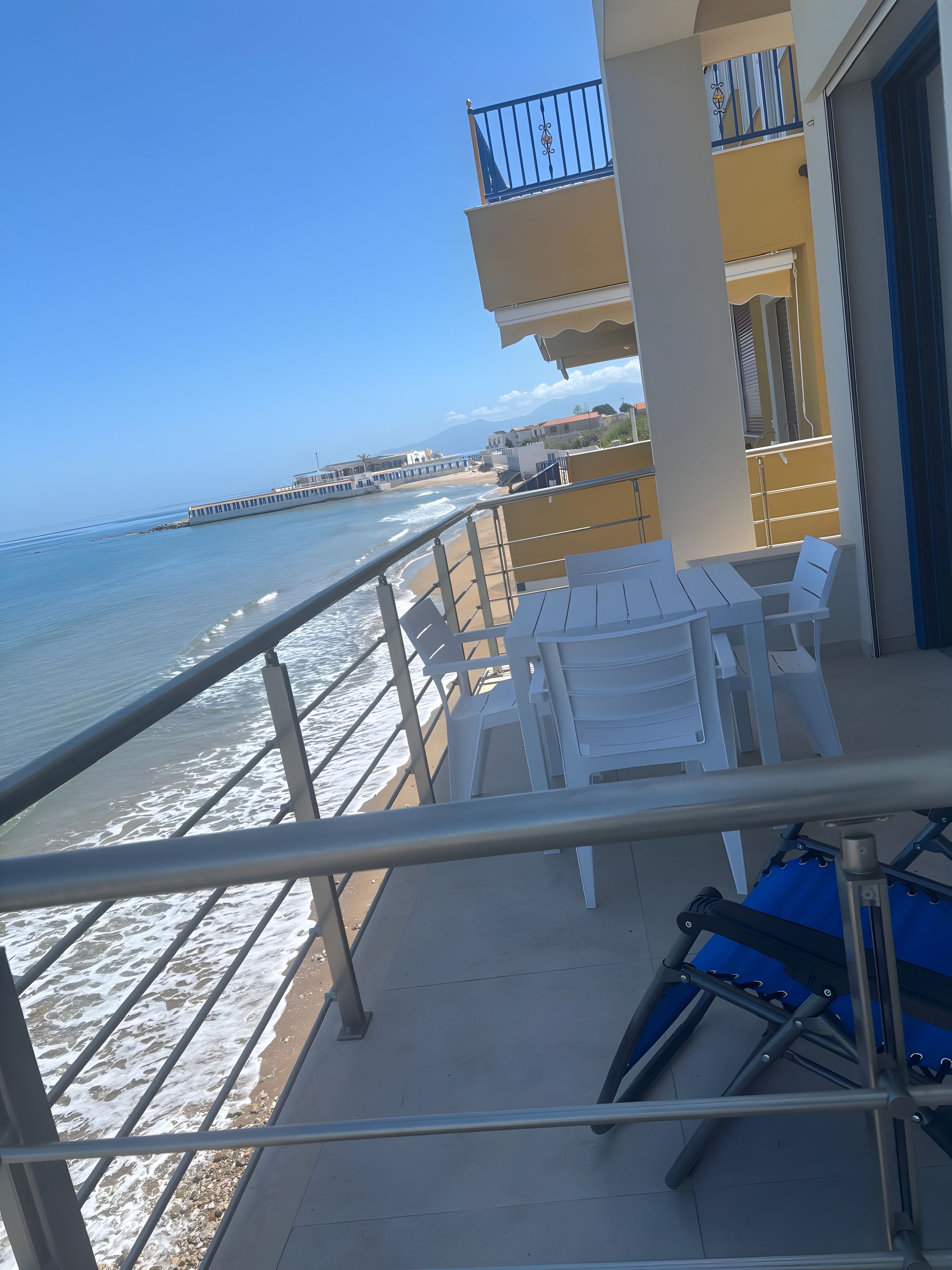 Un balcon avec vue sur la plage et l’océan, des chaises en plastique blanches et une chaise pliante bleue.