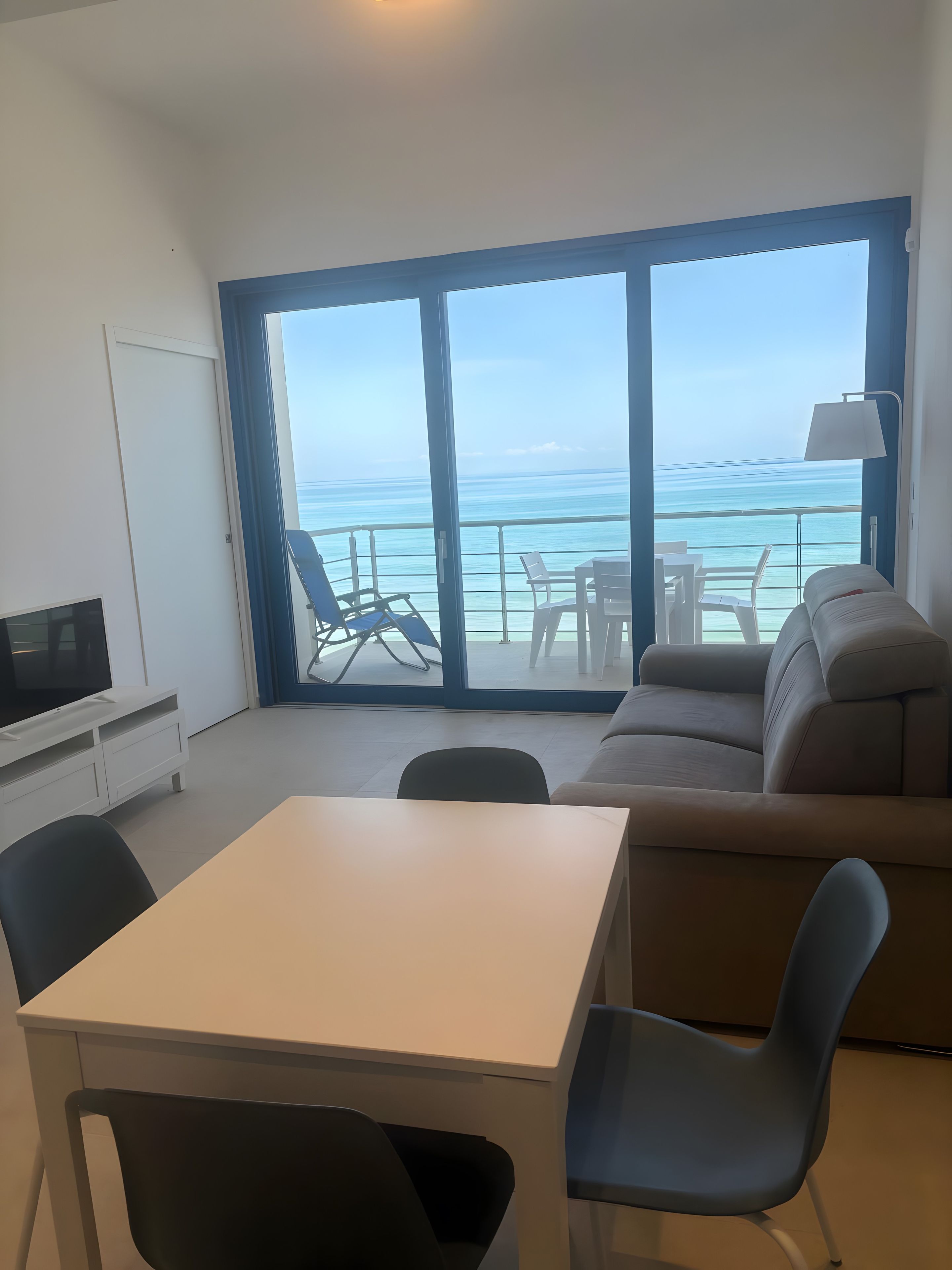 Une pièce avec une télévision, une table et un canapé. Il y a une grande fenêtre donnant sur la mer.