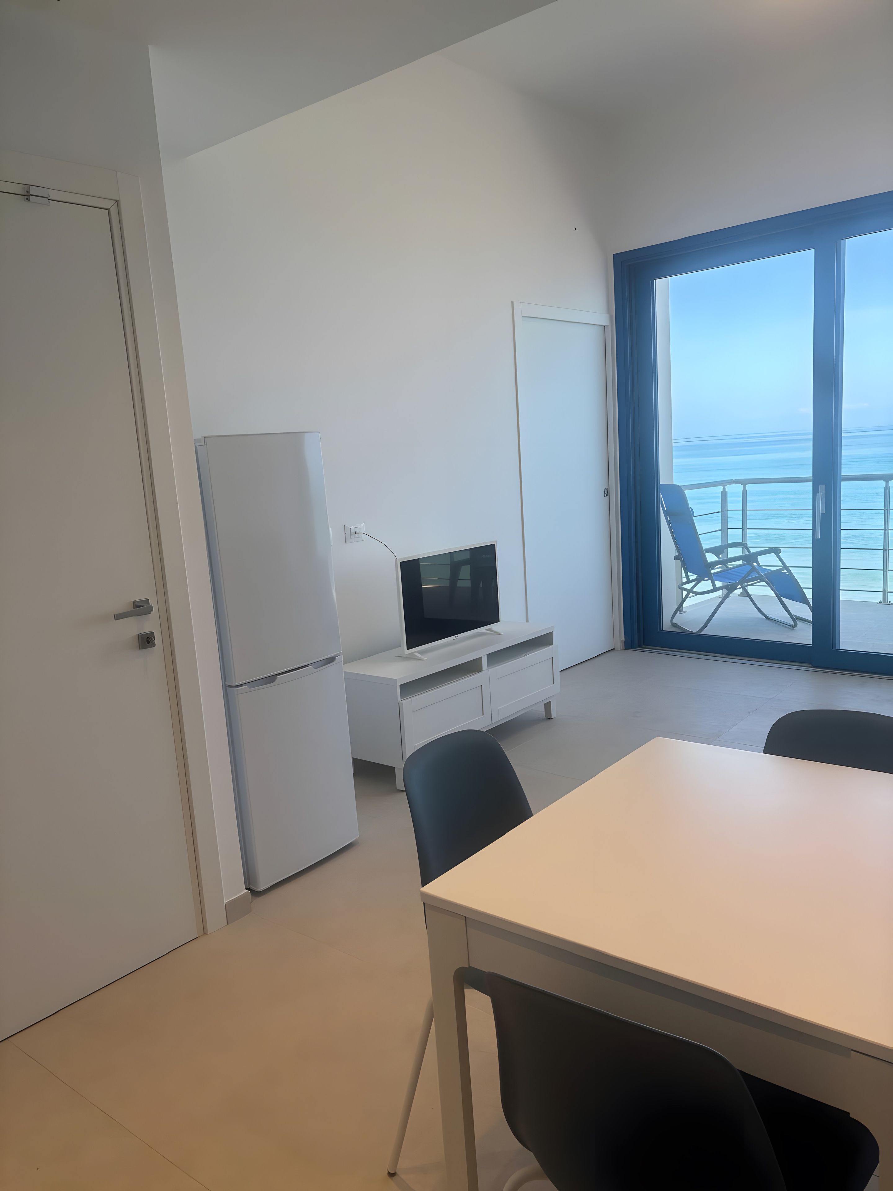 Una habitación con un escritorio blanco, una silla negra, un televisor sobre un mueble blanco y vista al océano a través de un ventanal grande.