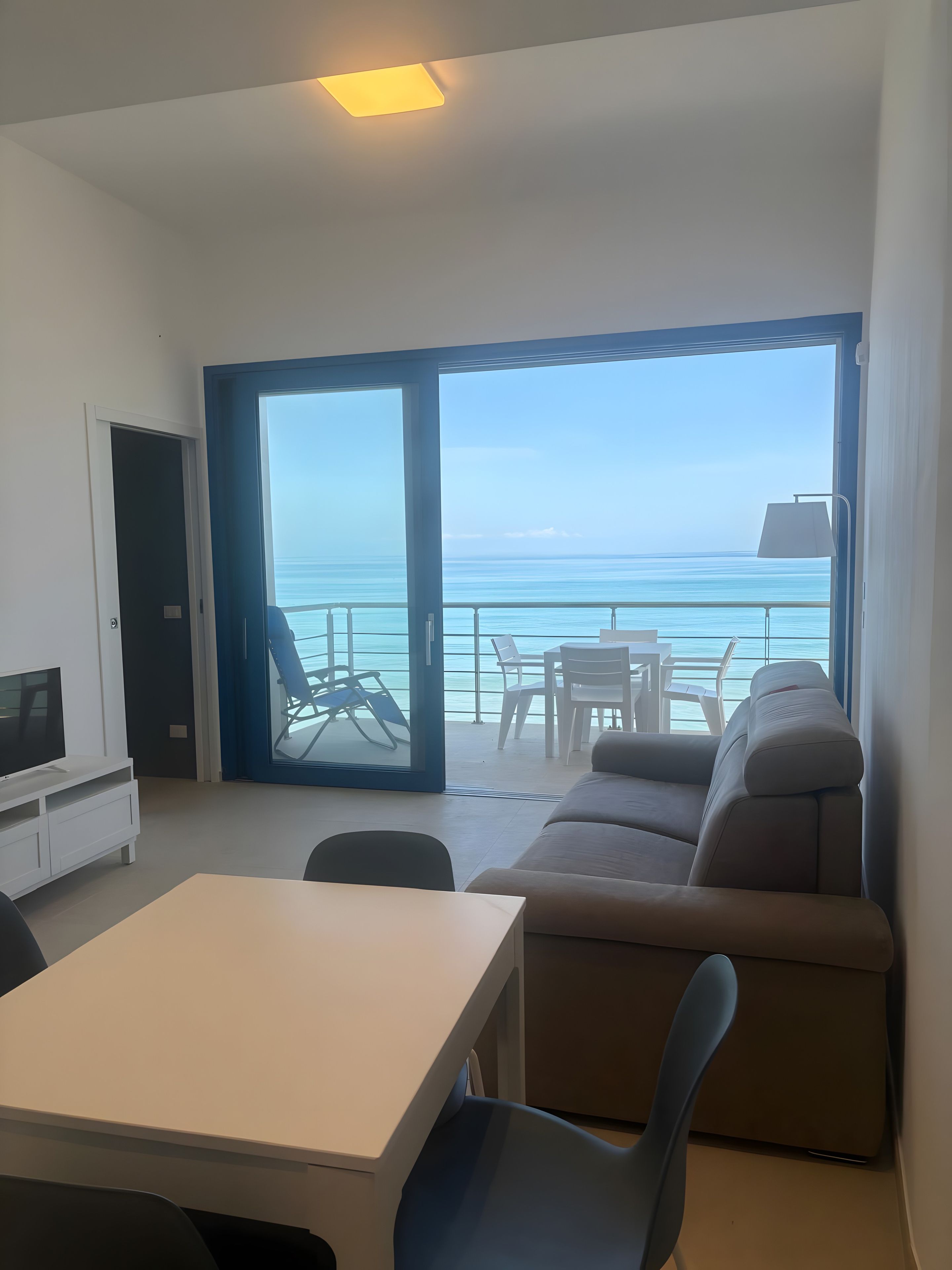 A room with a sofa, a table, and a TV. There is a door leading to a balcony with a view of the sea.
