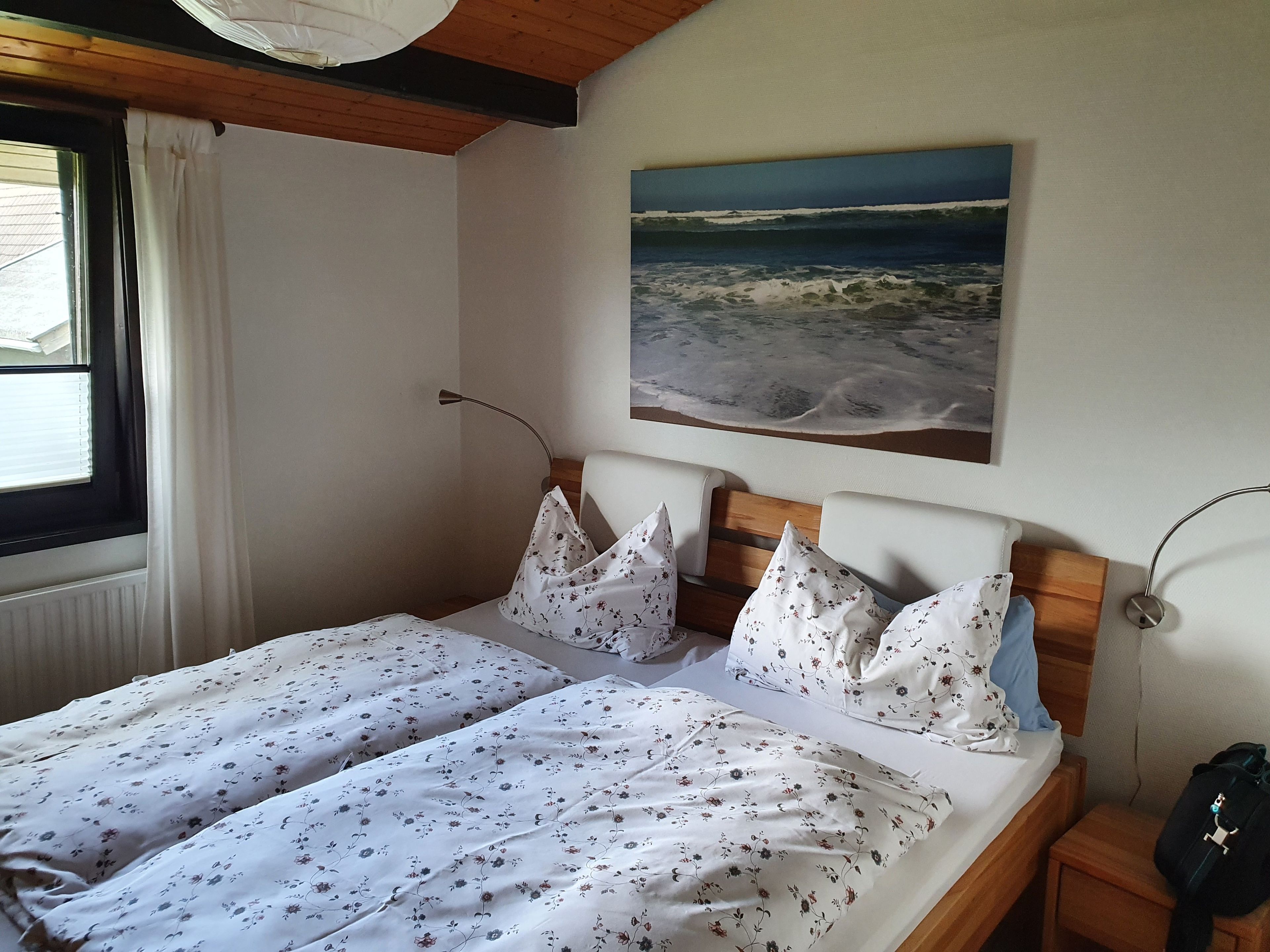 Ein Doppelbett mit floralem Bettzeug, ein Gemälde mit Strandmotiv und ein Fenster mit Vorhängen.