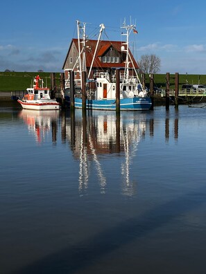 A calm river with two boats docked at a pier, a house with a red roof, and a clear blue sky. - Holiday Home '"Marschenhuus"' with Wi-Fi (Butjadingen)