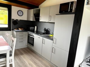 A kitchen with a microwave, oven, and sink. There is a clock on the wall and a window with a view of greenery. - Holiday Home '"Marschenhuus"' with Wi-Fi (Butjadingen)