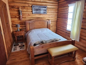 Ein Schlafzimmer einer Holzhütte mit Bett, Bank, Nachttisch, Lampe und einem gerahmten Bild an der Wand.
