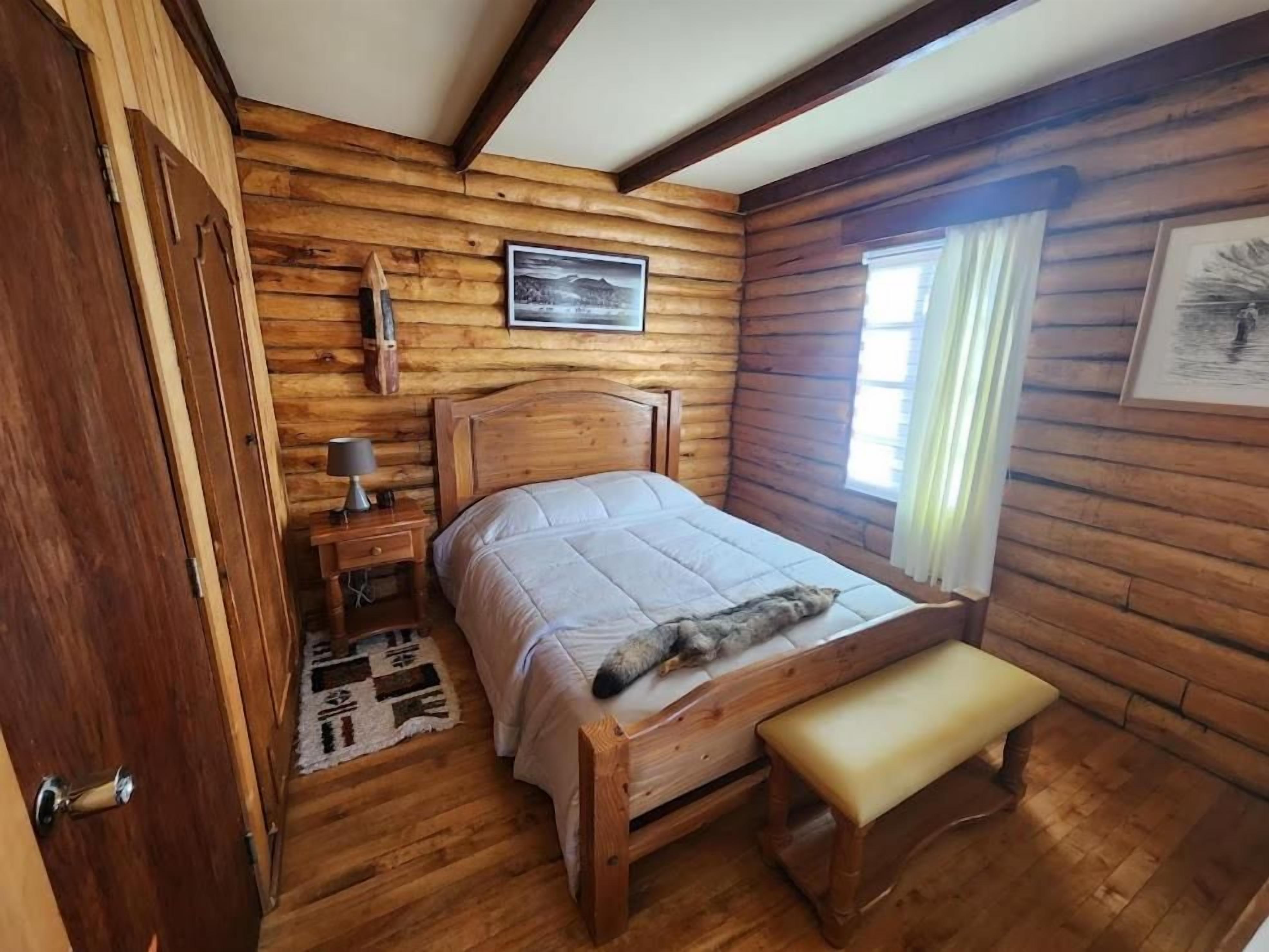A wooden cabin bedroom with a bed, bench, bedside table, and a window with curtains.