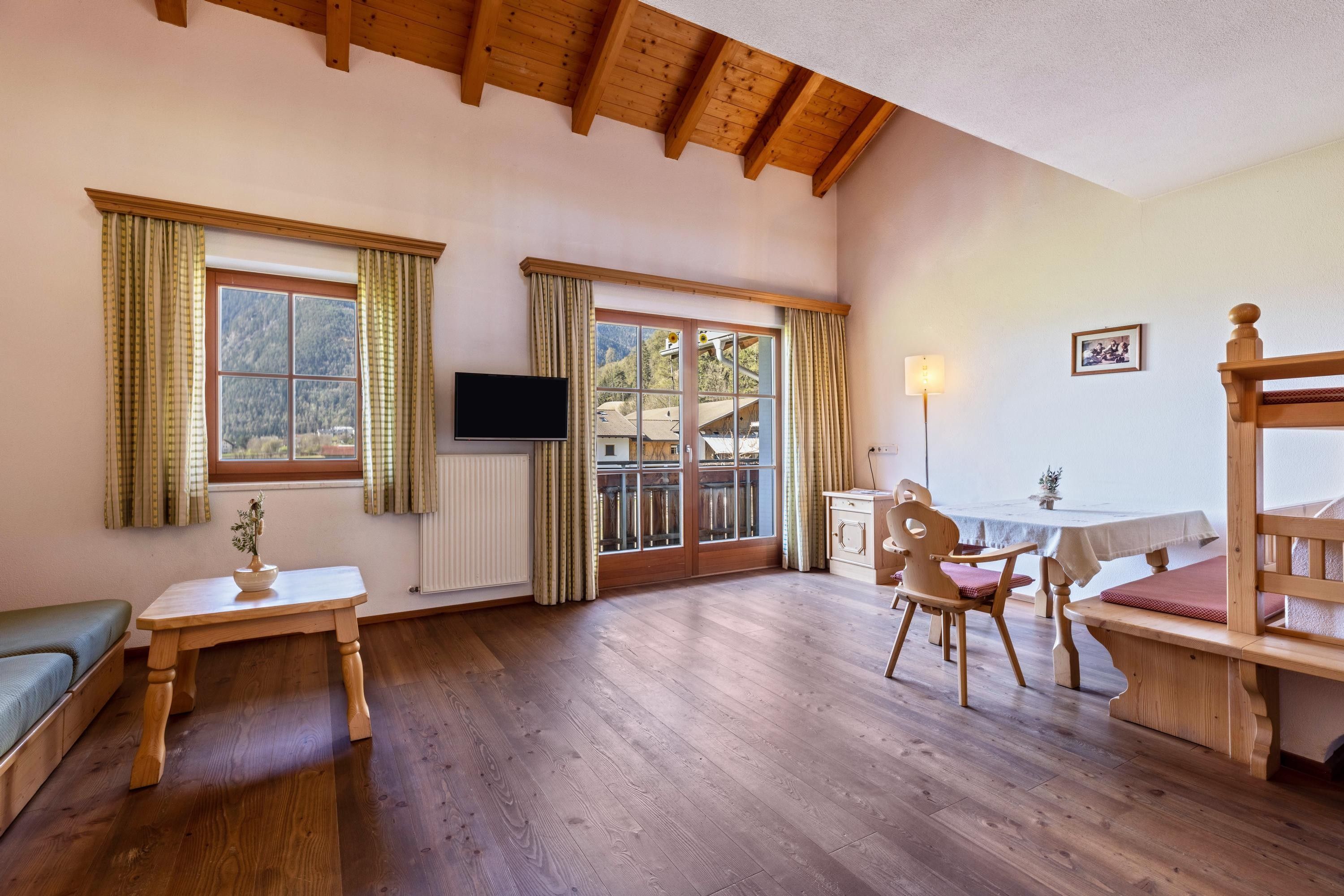 Une pièce avec un plancher en bois, une télévision, une table, des chaises et une fenêtre avec des rideaux.