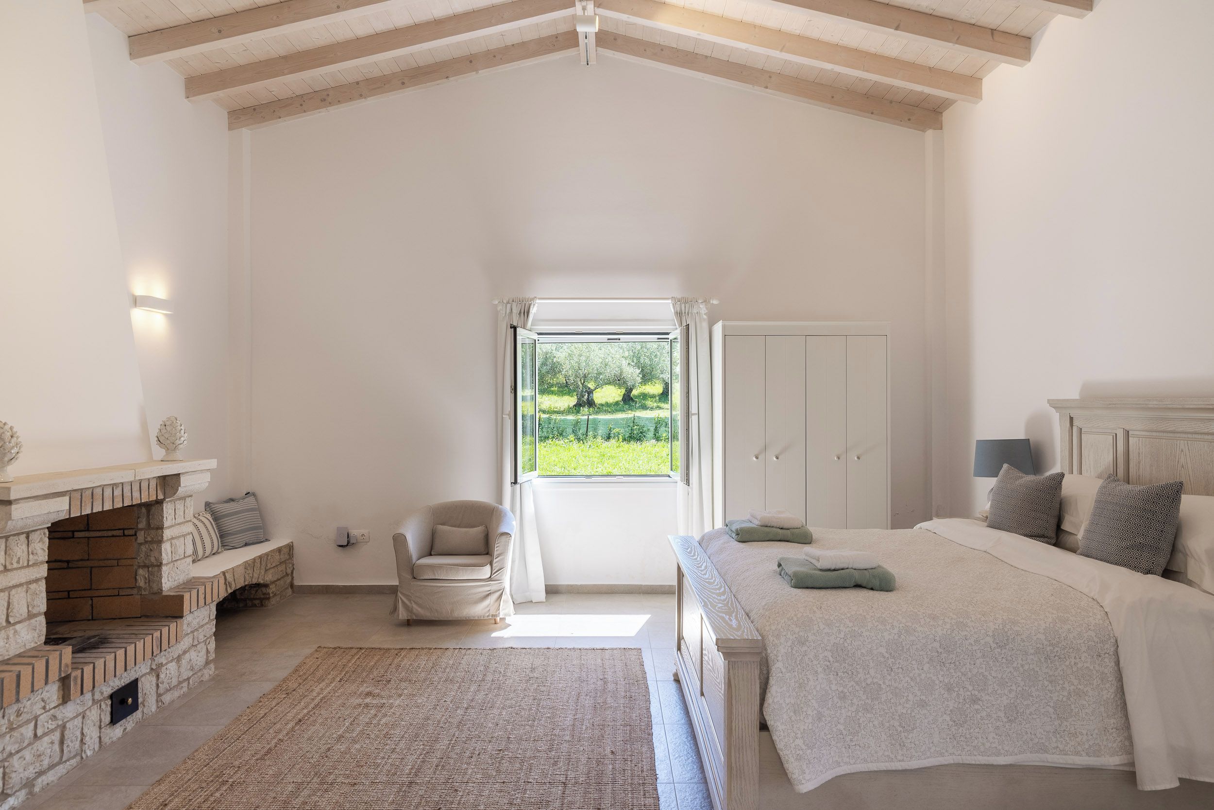 A bedroom with a large bed, a chair, a fireplace, and a view of a garden through a window.