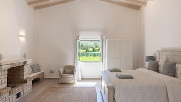 A bedroom with a large bed, a chair, a fireplace, and a view of a garden through a window.