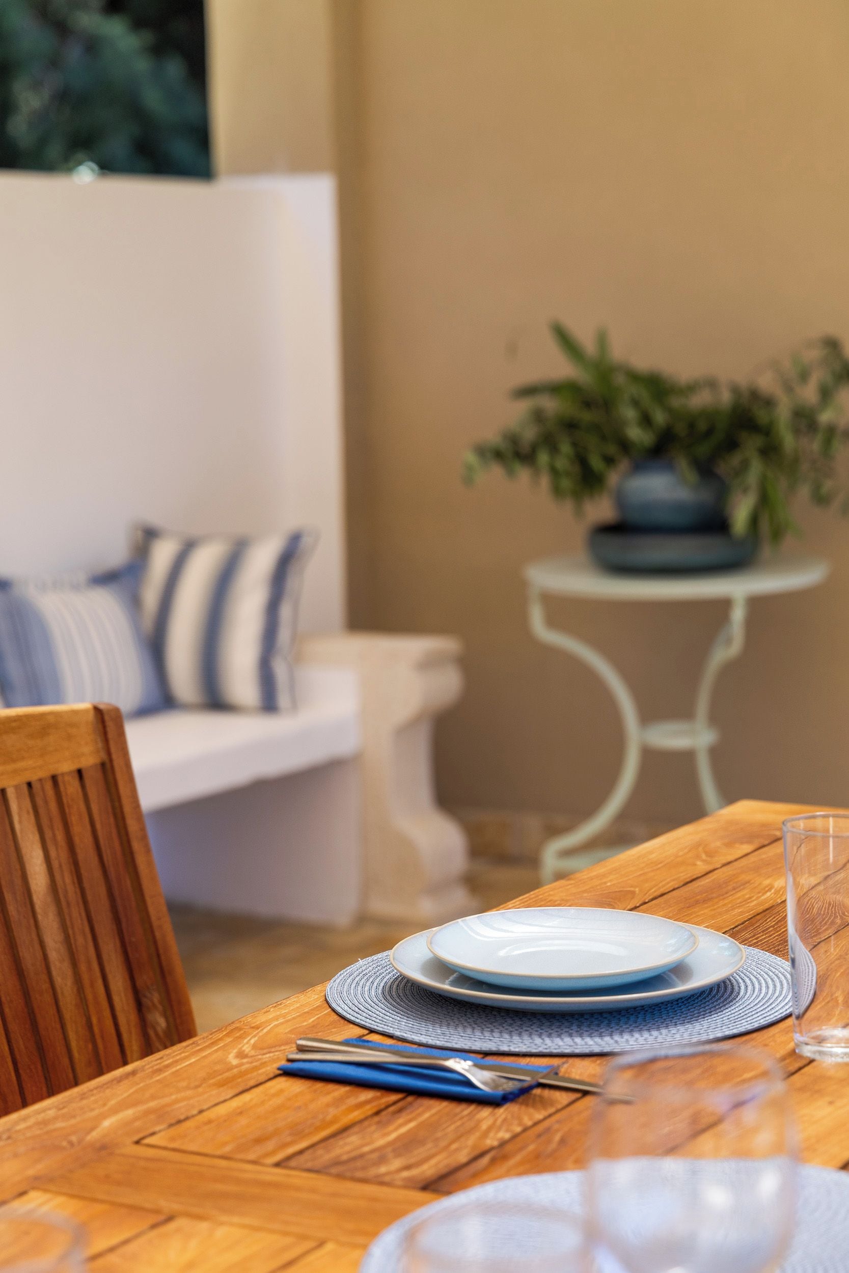 Mesa de comedor dispuesta con platos, cubiertos y una copa. Hay una silla frente a la mesa, una mesita con una planta y un banco blanco con cojines a rayas al fondo.