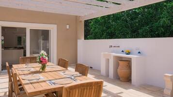 An outdoor patio with a wooden table set for dining, surrounded by chairs, and a white wall with a built-in grill and decorative items.
