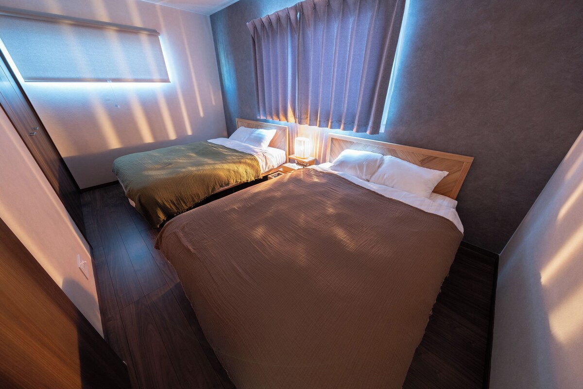 Quarto de hotel com duas camas, cabeceira de madeira, mesa de cabeceira com abajur e janela com cortinas.