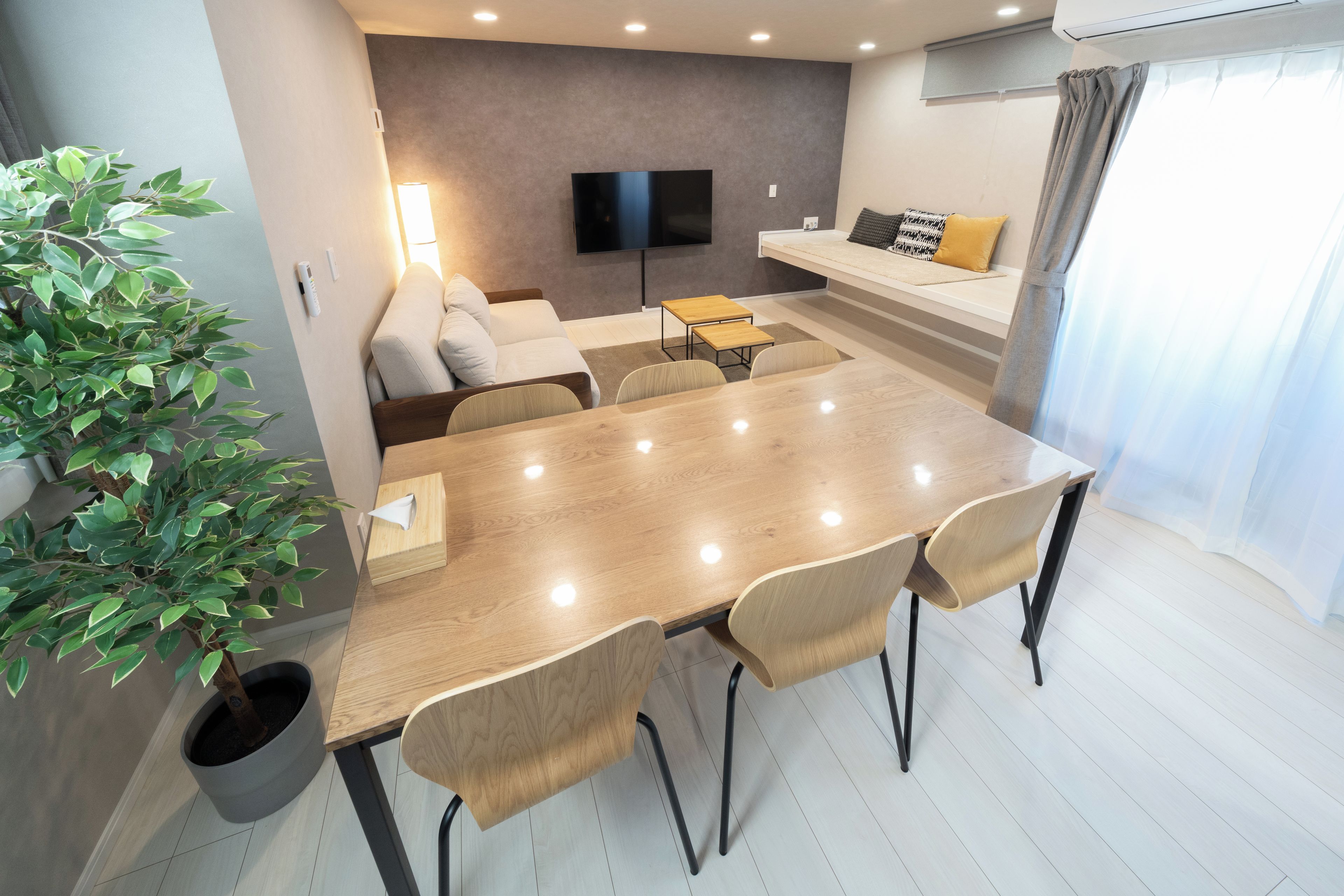 A modern living room with a flat-screen TV, a sofa, a dining table with chairs, and a potted plant.