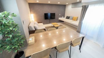 A modern living room with a flat-screen TV, a sofa, a dining table with chairs, and a potted plant.