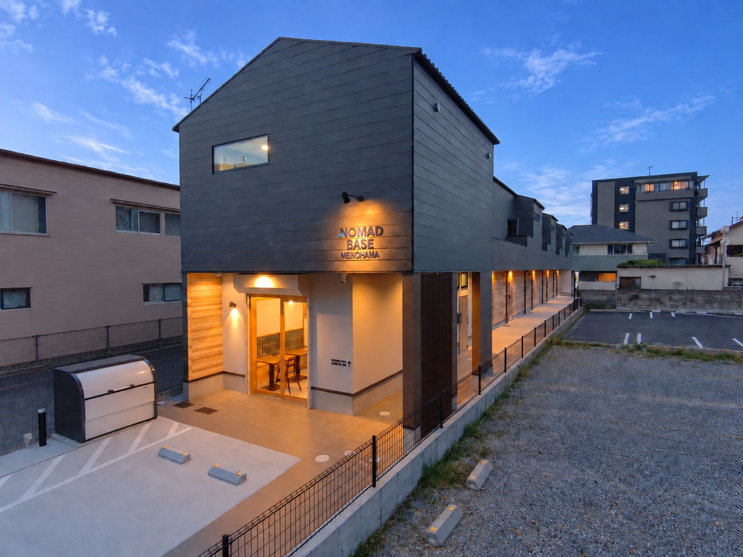 Un edificio moderno con un letrero que dice 'NOMAD BASE MEINOHAMA' y un área de estar al aire libre techada.