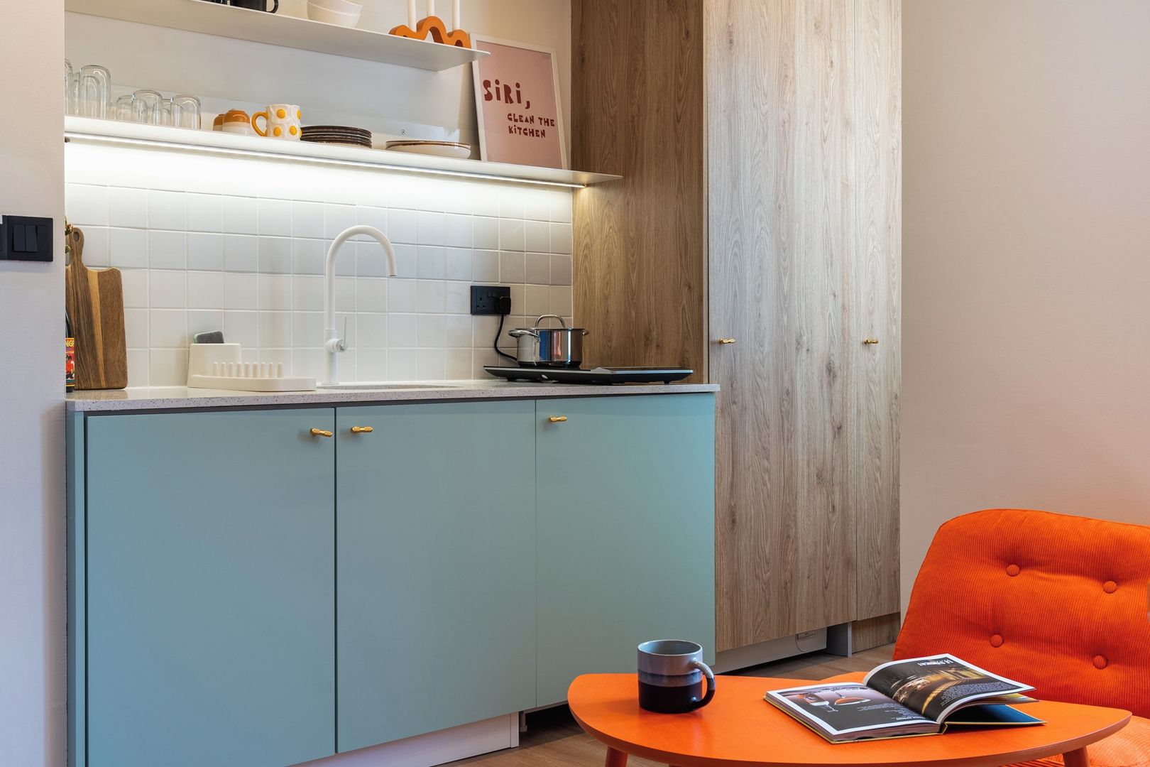 Une cuisine avec des meubles bleu clair, un plan de travail blanc et un mur en bois. On y trouve une petite table orange sur laquelle sont posés un livre et une tasse.
