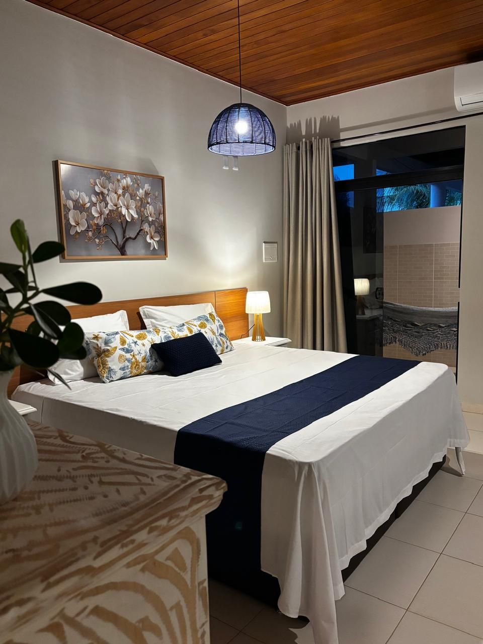 A hotel room with a bed, bedside table, lamp, and a view of the outside.